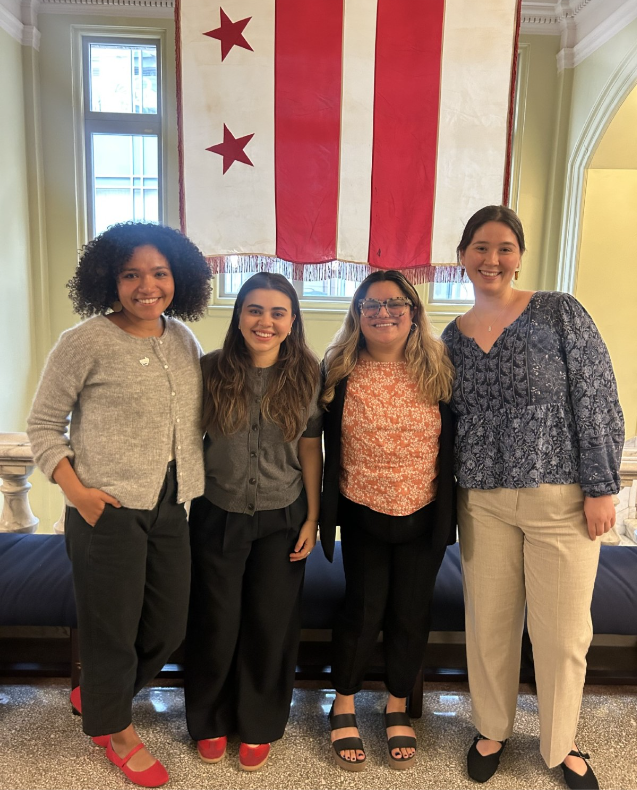 Four Ayuda staff members in professional dress pose before the District of Columbia flag at the Wilson Building.
