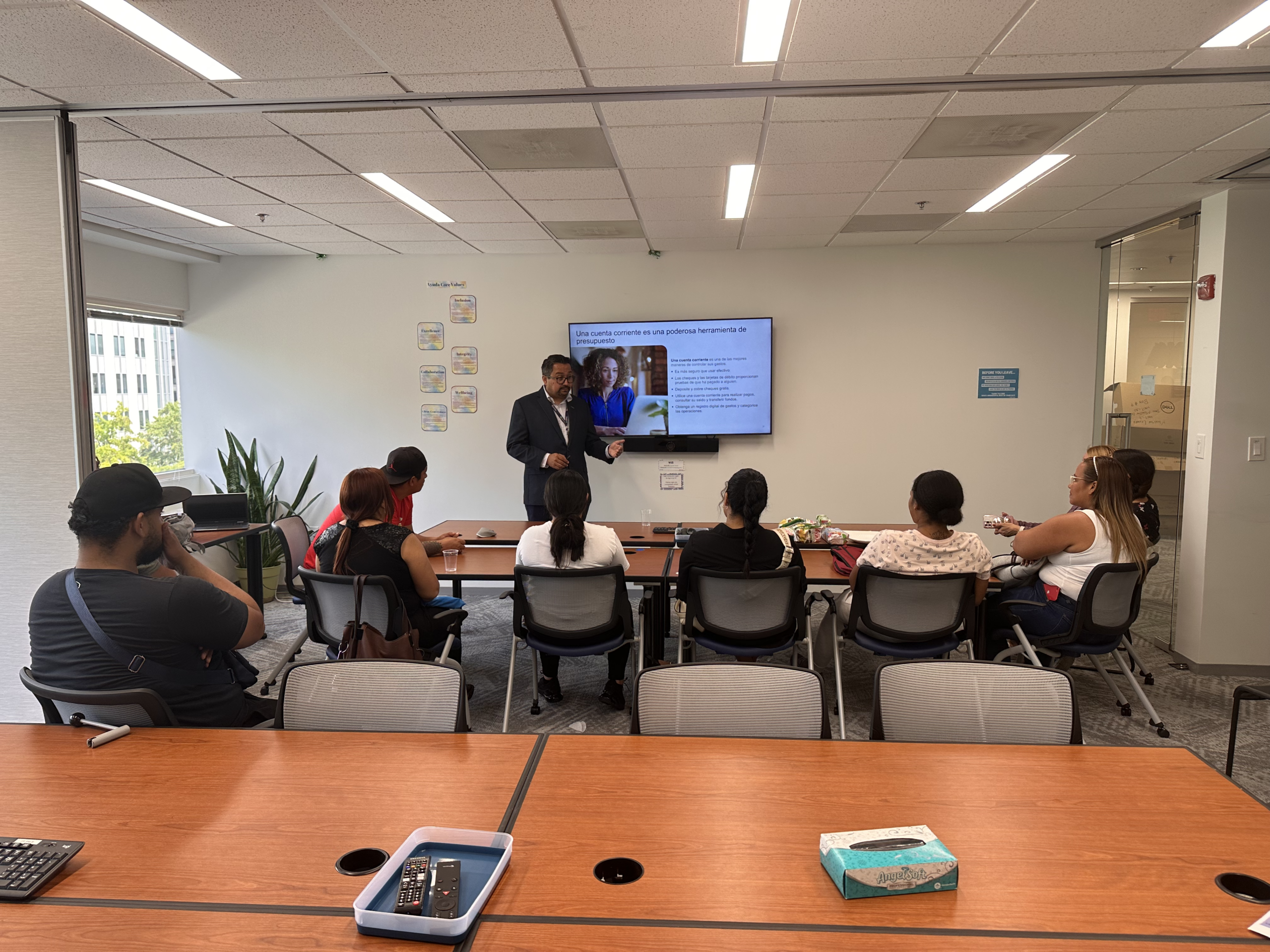 An instructor leads a financial literacy workshop for Ayuda clients seated around a conference room table, with a presentation about budgeting displayed on a screen.