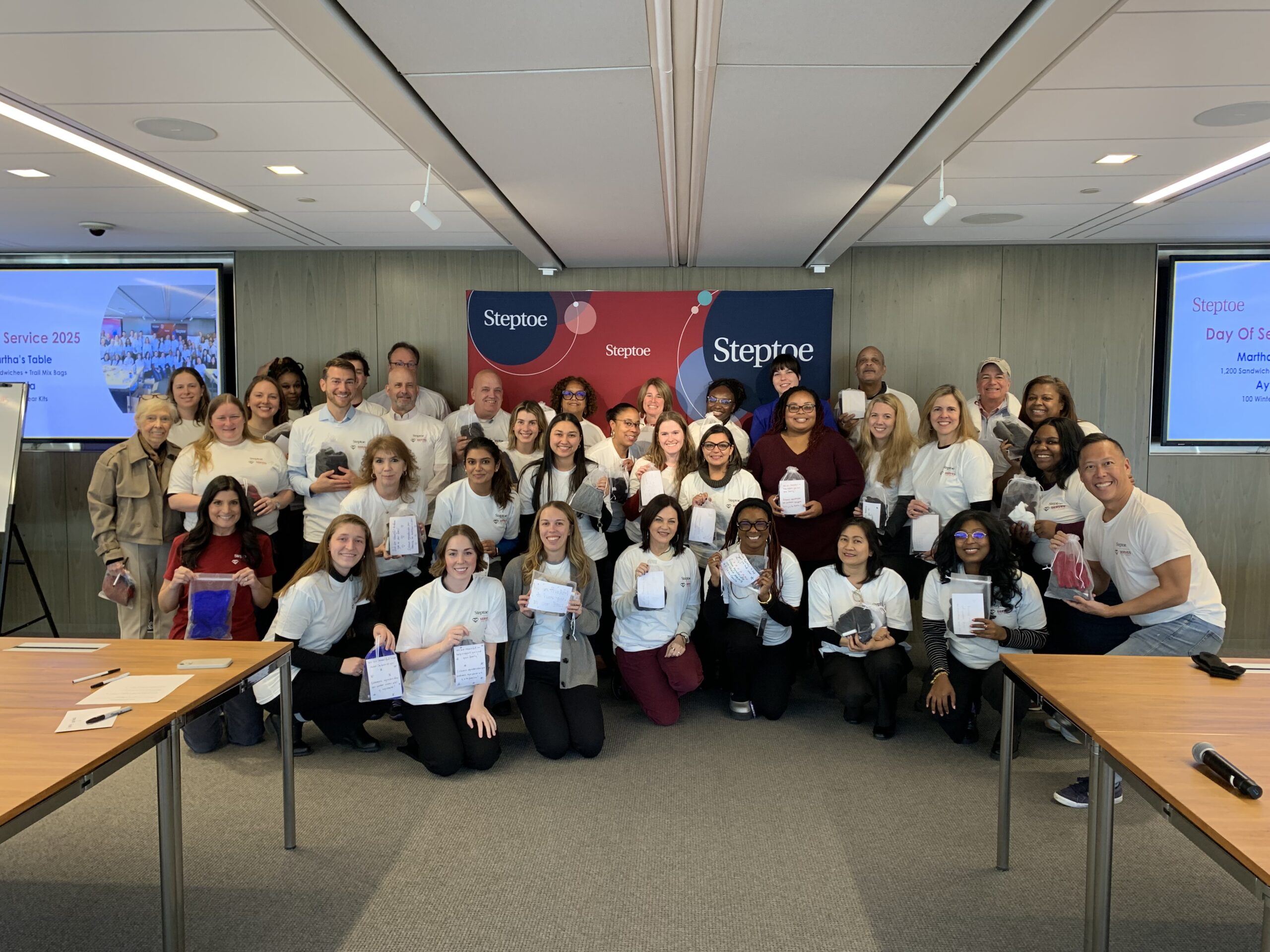 A large group of Steptoe volunteers and Ayuda team members pose in a conference room while holding assembled winter care packs for clients.