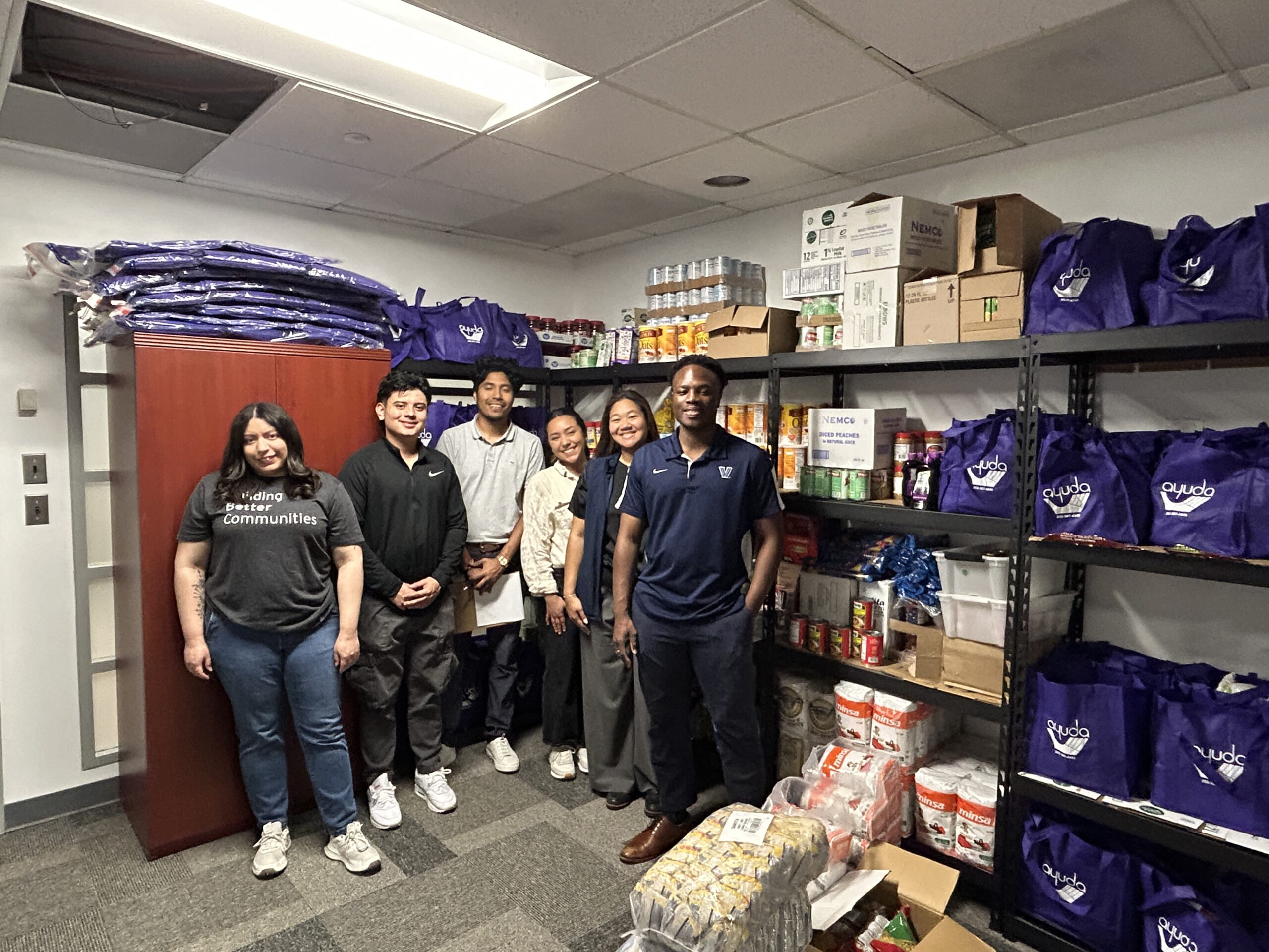 Six volunteers stand in Ayuda’s food pantry surrounded by shelves stocked with canned goods, pantry staples, and purple Ayuda bags prepared for community distribution.
