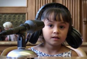 A very young girl sits in a courtroom, wearing a headset that's falling off because of how big it is. 